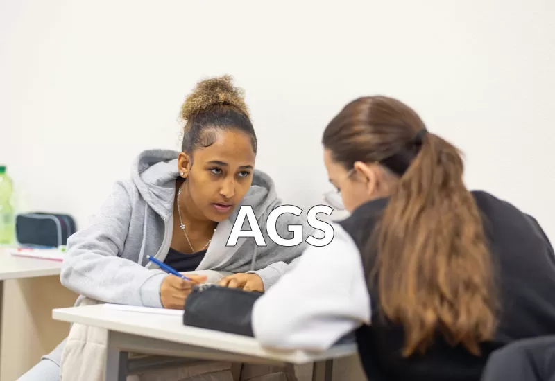 Zwei AGS-Lernende am Schultisch: eine schreibt, die andere hört aufmerksam zu.