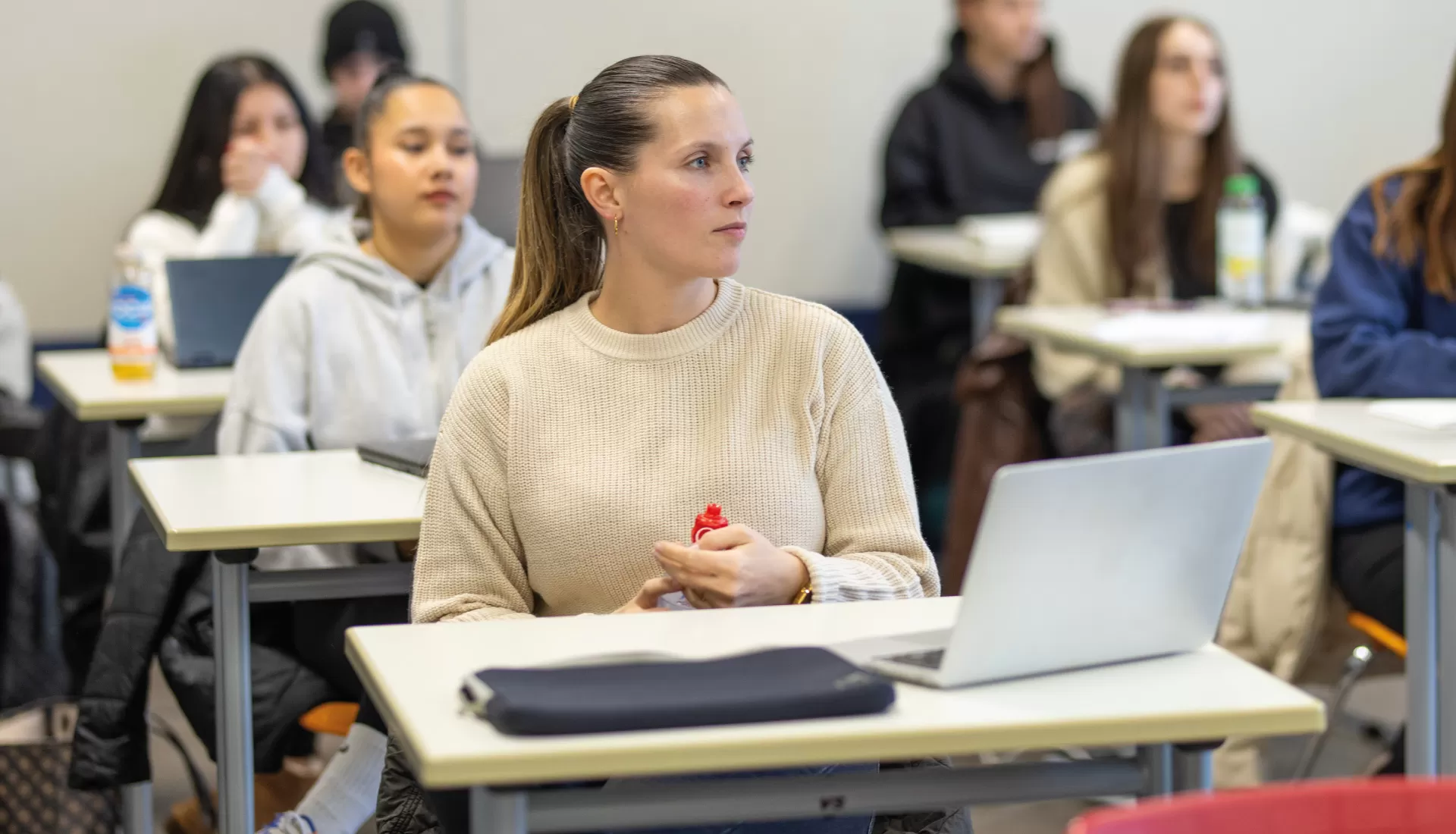 Frau sitzt aufmerksam in einem Klassenzimmer an einem Tisch mit Laptop, im Hintergrund weitere Lernende.
