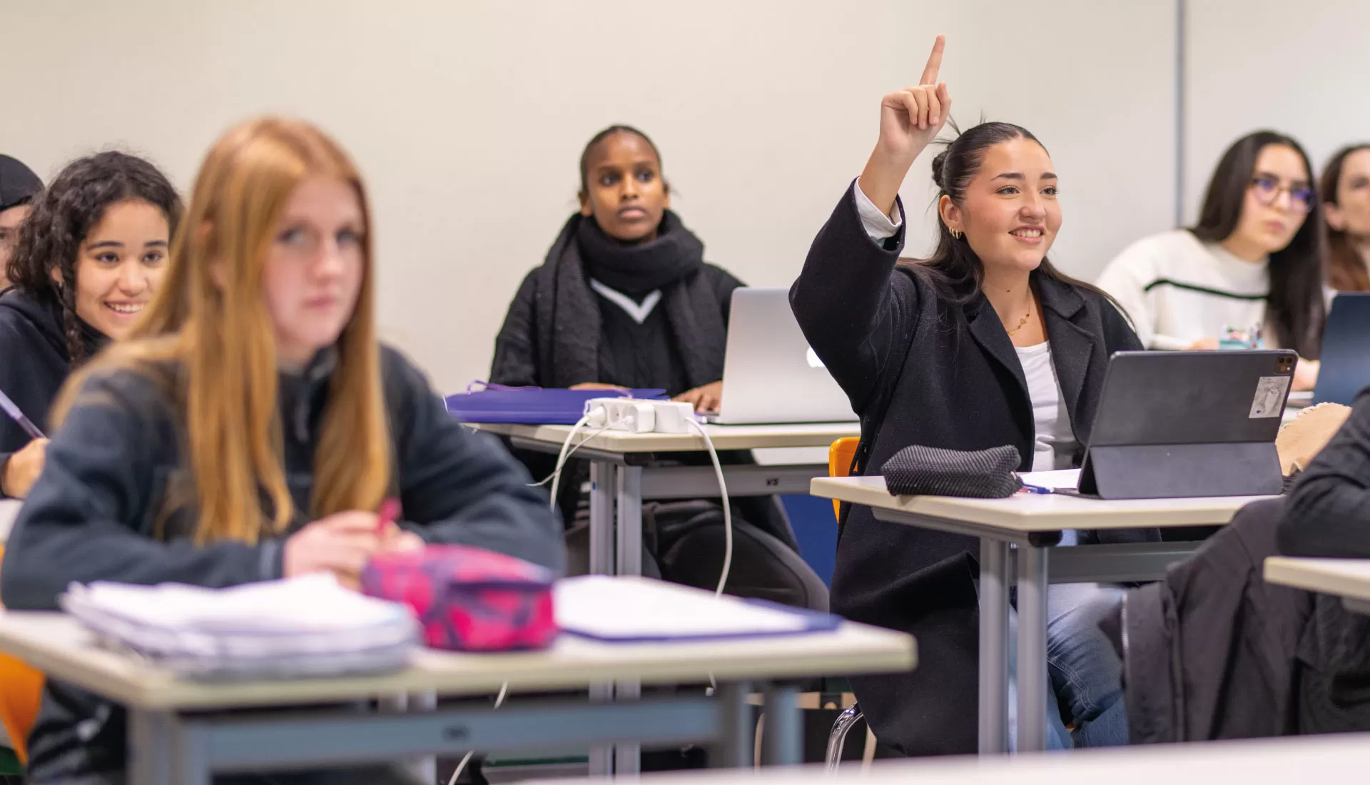 Lernende im Klassenzimmer; eine Person hebt lächelnd die Hand, um sich am Unterricht zu beteiligen.