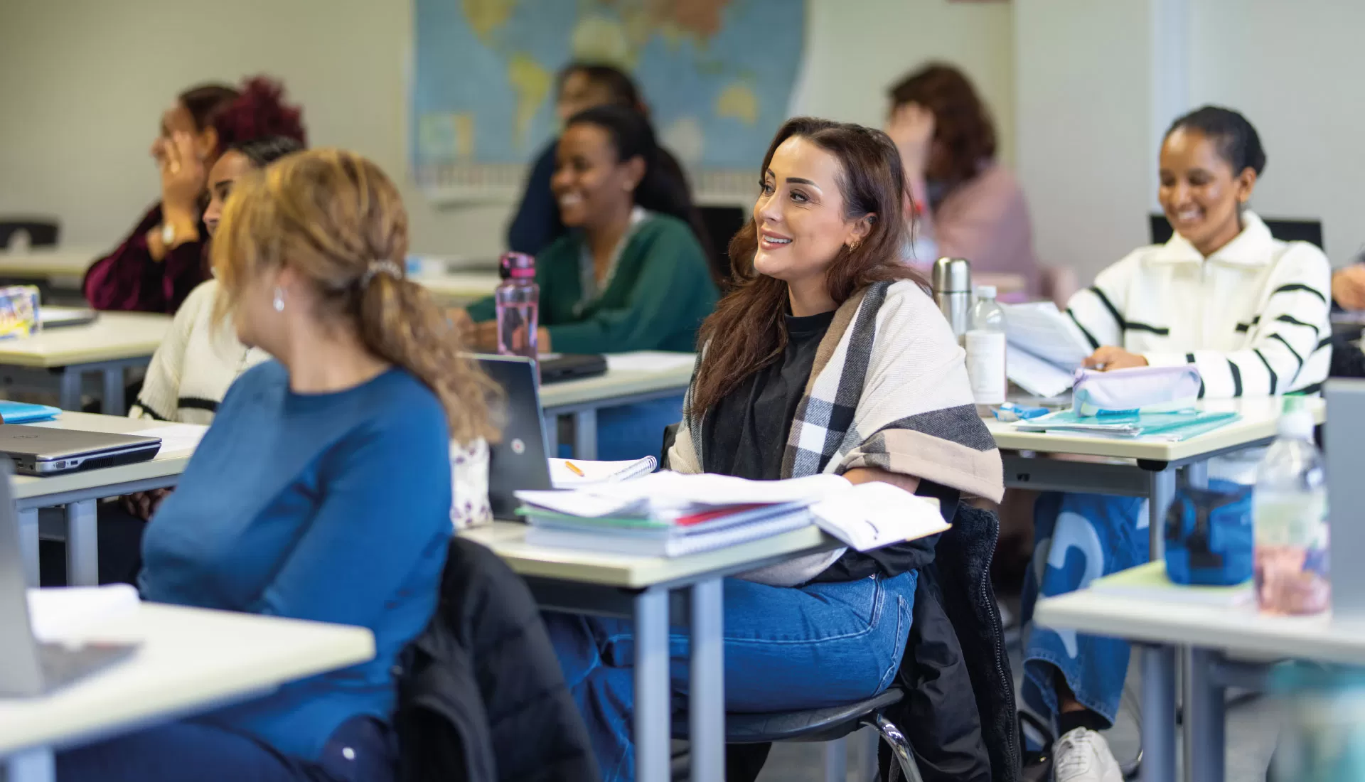 Lernende sitzen mit Unterlagen und Laptops im Klassenzimmer und hören aufmerksam zu.