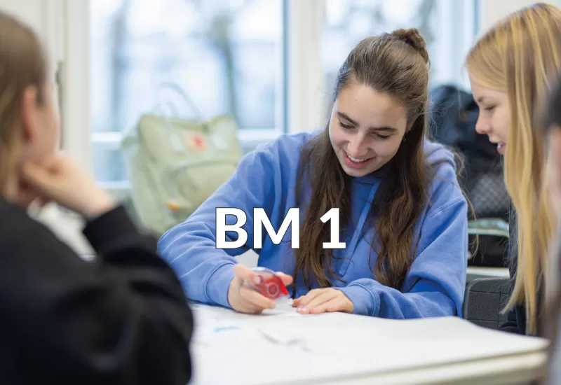 Lernende arbeiten gemeinsam im Klassenzimmer, eine Schülerin schreibt lachend etwas auf.