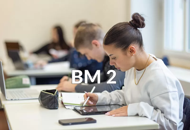 Schülerin schreibt mit Tablet-Stift, weitere Lernende im Hintergrund im Klassenzimmer.